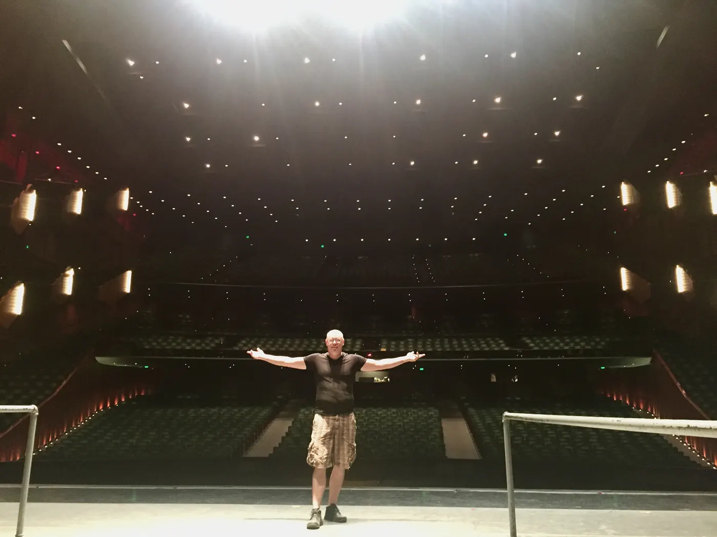 Standing alone on a large stage, arms out, facing an empty auditorium